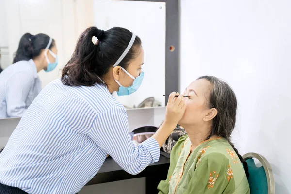 在化妆室里 女性化妆师的照片用眼影罩在她年老的客户脸上 — 图库照片