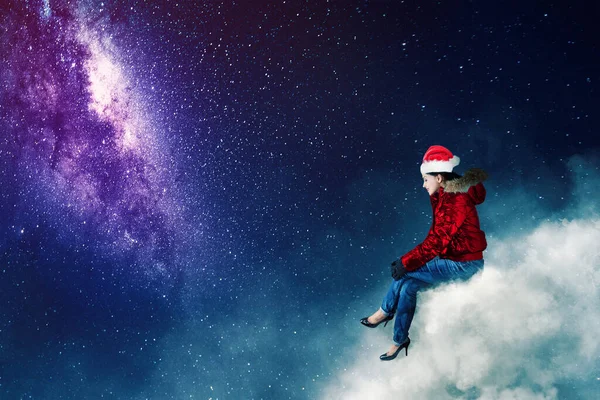年轻女子坐在云彩上 望着夜空中的银河 头戴圣诞礼帽 身穿毛衣 — 图库照片