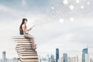 Kadın üniversite öğrencisi, modern şehir arka planına sahip kitapların üzerinde otururken parlak parçacıkların ışıklarıyla kitap okuyor.