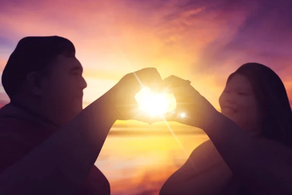 在日落时站在海滩上用手做心脏标志的一对年轻肥胖夫妇的亲密接触 — 图库照片