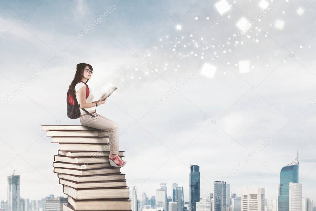 Female college student reading a book with bright particles lights while sitting on heaped books with modern city background