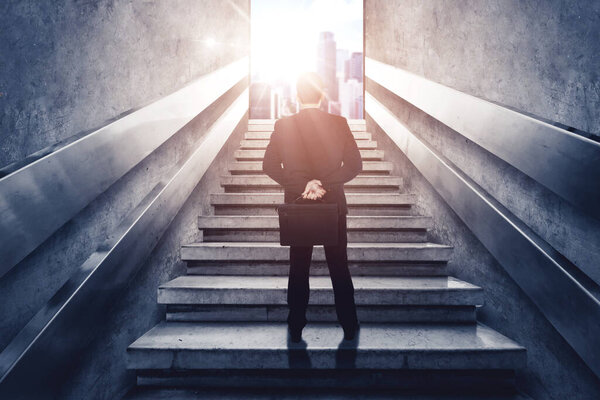 Rear view of male manager holding a suitcase while standing on the stairs and looking at bright modern city