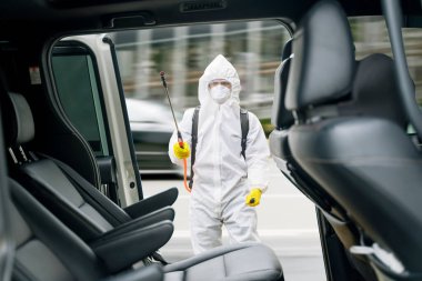 Tehlikeli madde giysisi giyen ve arabanın içini spreyleyen erkek işçi, toplu taşımada koronavirüsün yayılmasını önlüyor