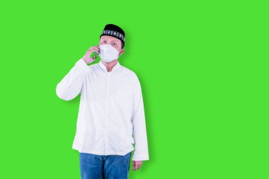 Arama yaparken yüz maskesi takan Müslüman genç bir adam stüdyoda duruyor ve yeşil ekran arka planı var.