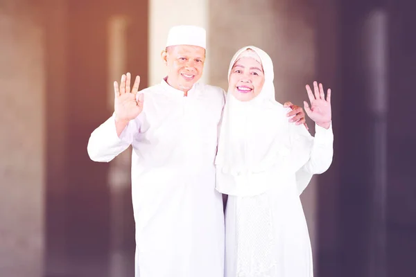 Smiling Muslim senior couple waving hands at the camera and standing together in the mosque