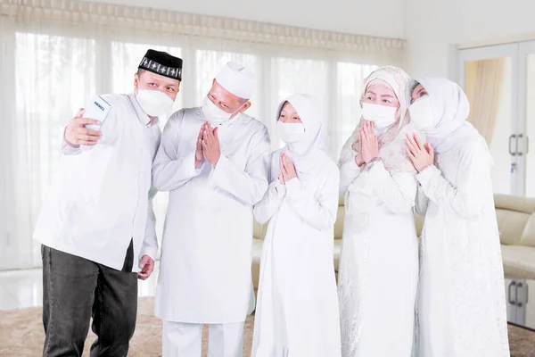 Three generation Muslim family wearing face mask while showing congratulate hands gesture Eid Mubarak at cellphone during video call in the living room at home