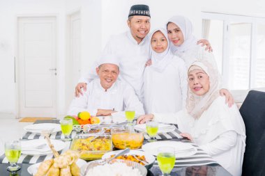 Üç nesildir neşeli Müslüman bir aile birbirlerine sarılıyor ve evde bayram yemeği yerken kameraya gülümsüyor.