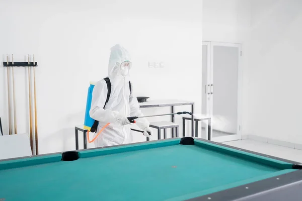 コロナウイルスのパンデミック時に消毒スプレーでビリヤード台を掃除しながらハズマットスーツを着た若者 — ストック写真