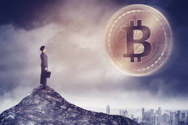 Modern şehrin üstündeki bulutlu gökyüzünde altın bir bitcoin 'e bakarken ve tepelerde dururken evrak çantası taşıyan bir kadın yönetici.