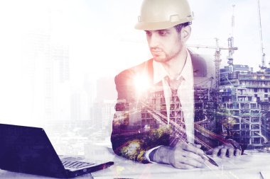 İnşaat alanının arka planında otururken dizüstü bilgisayar ve planlarla çalışan erkek müteahhitlerin iki katına çıkarılması