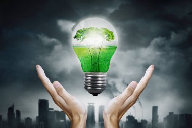 Enerji kaynağı kavramı. Şehrin kirli arka planına sahip ampulün içinde yeşil doğayı tutan bilinmeyen ellere yakın durun.