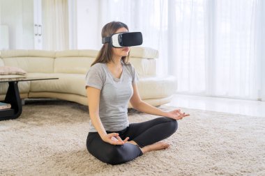 Slim woman doing yoga exercise and meditation with a Virtual Reality glasses in the living room. Shot at home