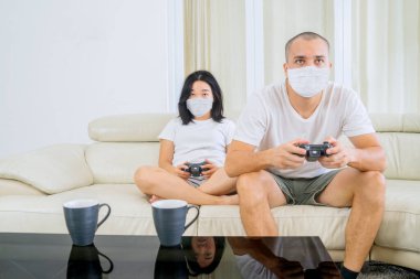 Young couple playing video games together at home while wearing face mask during self isolation in the living room