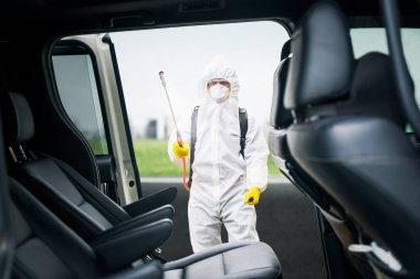 Young man wearing protective suit while spraying disinfectant to car interior on the roadside for preventing coronavirus transmission