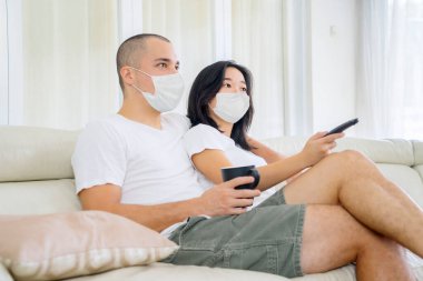 Young couple watching television together at home while wearing face mask during self isolation in the living room