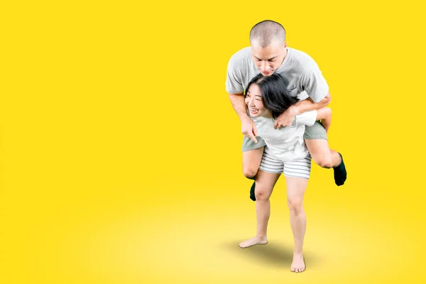 Strong young woman looks happy while giving her boyfriend piggyback ride in the studio with yellow background