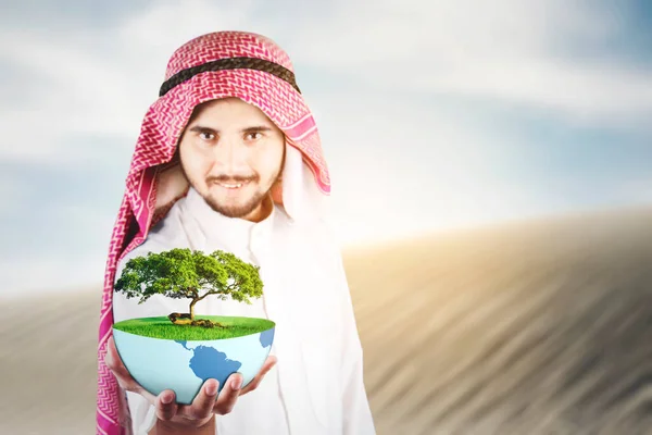 Arabian man holding an earth planet and green tree while standing in the desert