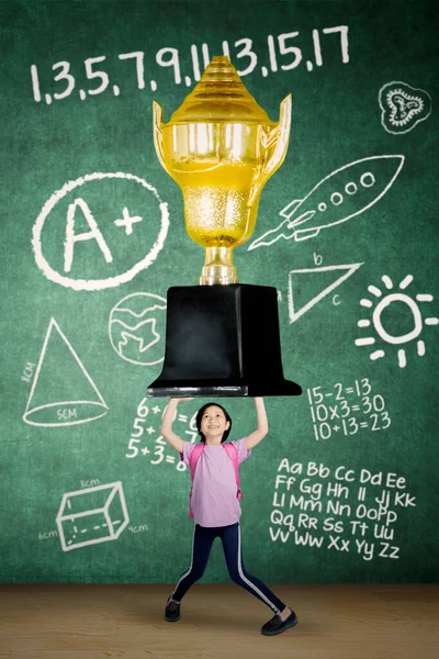 快乐的女学生一边提着一个大奖杯 一边站在教室里背负着涂鸦 — 图库照片