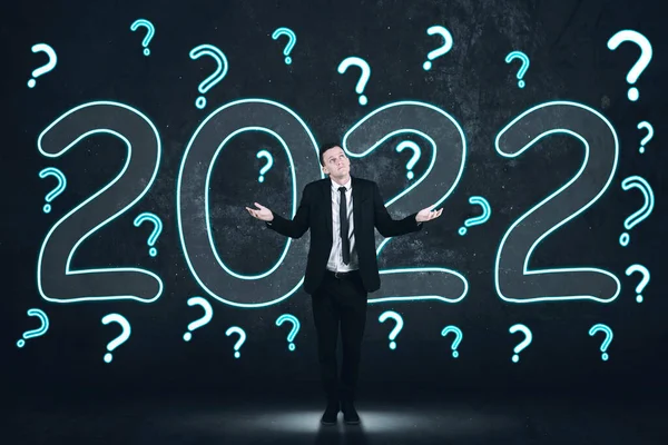 Confused businessman shrugging hands while standing with question marks and 2022 numbers in dark background