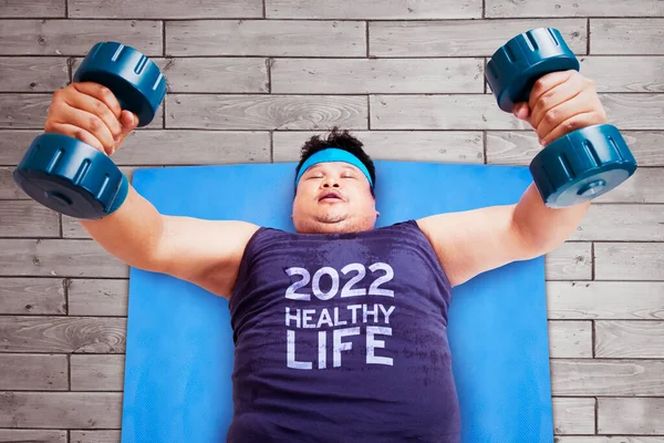 彼のシャツの上に2022の健康的な生活のテキストと横になっている間 2つのバーベルで運動脂肪男のトップビュー — ストック写真