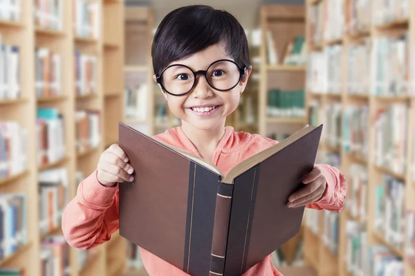 Schönes Kind mit Brille, das Buch in der Bibliothek hält — Stockfoto