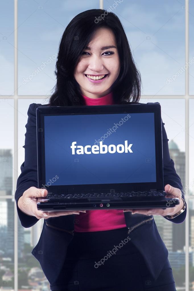 JAKARTA, SEPTEMBER 08, 2015: Image of beautiful businesswoman smiling at the camera while showing laptop computer with facebook logo