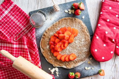 Çilekli ve bademli ev yapımı turtalı kek, siyah masa ve ahşap masa. Yanında yemek aletleri olan kırmızı havlu.