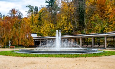 Luhacovice, Çek Cumhuriyeti 'ndeki spa meydanının sonbahar manzarası, merkezinde bir çeşme ve arka planında kırmızı, turuncu ve sarı tonlarda parlayan renkli ağaçlar var..