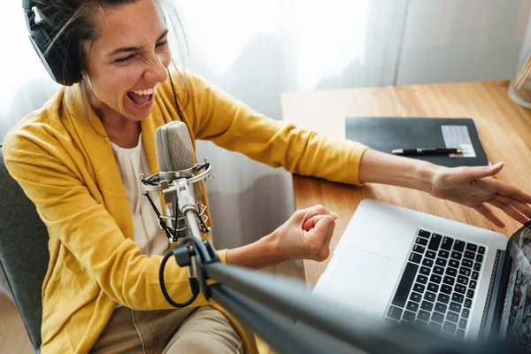 Charismatic woman radio host recording podcast at home studio. Female podcaster recording broadcasting into microphone using laptop