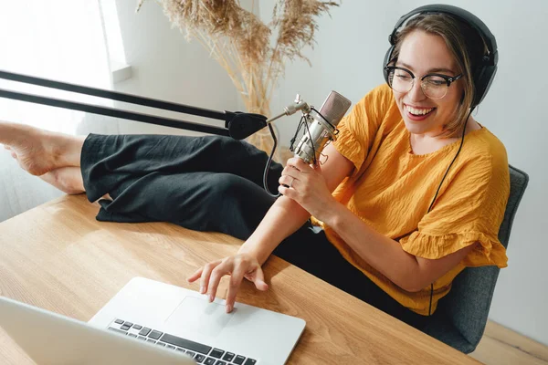 Charismatic woman radio host recording podcast at home studio. Female podcaster recording broadcasting into microphone using laptop