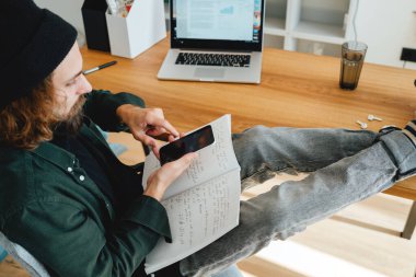 Sakallı öğrenci, dizüstü bilgisayar ve akıllı telefon kullanarak internetten ders çalışırken ayakları masanın üzerinde oturur. Üniversite öğrencisi üniversite dersine hazırlan.