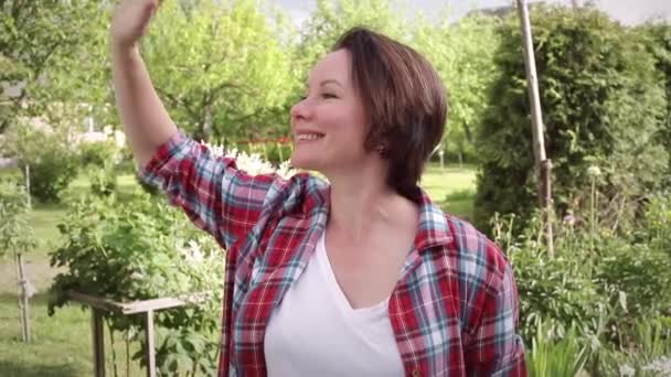 Una mujer con una camisa roja a cuadros saluda a su amiga. Escena callejera en el jardín — Vídeo de stock