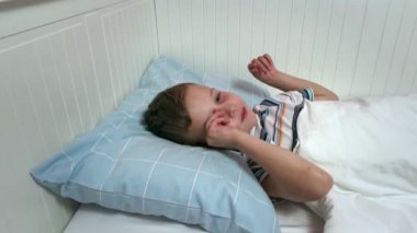 Çocuk sabah yatağında uyanır, gülümser ve saklanır. Mavi yatak, gözlerinin içine bak.