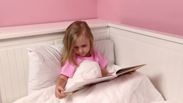 Une petite fille lit un conte de fées au lit avant d'aller se coucher. Livre à la main. Lit blanc — Video