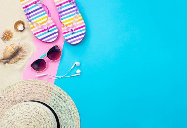 Metin için boş alanı olan modaya uygun yaz arkaplanı. Yaz tatili ürünleri mavi ve pembe arka planda sergilenir. Yaz şapkası, güneş gözlüğü, kulaklık, parmak arası terlik, kum ve deniz kabuğu. Güzel bir bayrak.