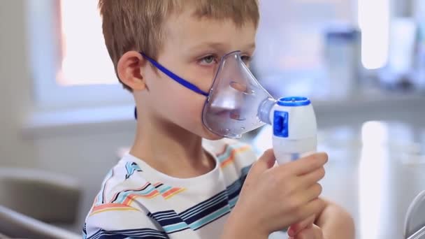 Un garçon de cinq ans portant un T-shirt à rayures bleues respire. Allergies, rhumes, toux, maladies respiratoires, nébuliseur. Dans la vidéo, l'enfant tient un masque de nébuliseur et regarde la caméra — Video