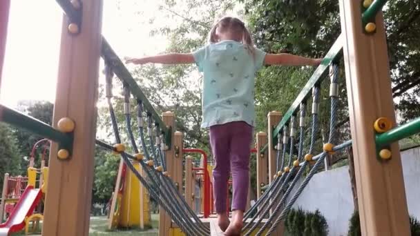 A girl walks with bare feet on a suspended log in an outdoor playground — Stock Video