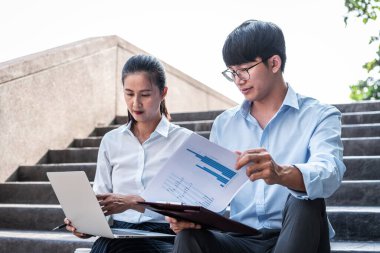 Genç iş adamı elinde iş kadınıyla dizüstü bilgisayardaki mali raporları kontrol etmek ve açık ofiste otururken iş projesi hakkında konuşmak için belge tutuyor.