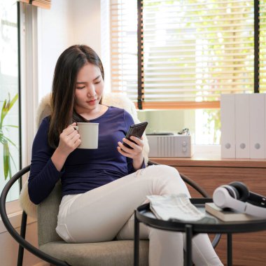 Genç Asyalı kadın elinde kahve fincanı tutarken ve akıllı telefondan video izlerken oturma odasındaki rahat koltukta dinleniyor.