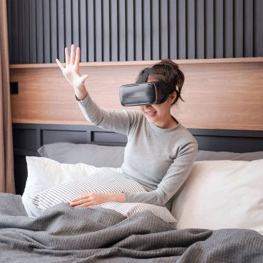 Genç Asyalı kadın, gündelik giysiler içinde havaya dokunmak için elini kaldırıyor ve dinlenirken sanal gerçeklik gözlüğü takıyor ve yatak odasında yatakta oturuyor.