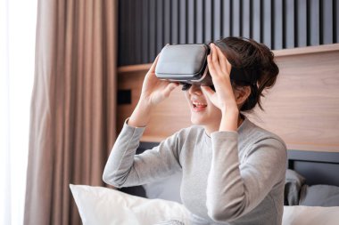 Genç Asyalı bir kadının günlük kıyafetlerini giyip oyun oynarken sanal gerçeklik gözlüğü takması dinlenmesi ve yatak odasında yatakta oturması.