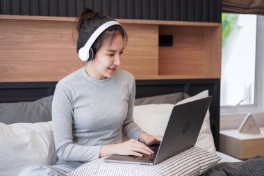 Kulaklıklı genç Asyalı bir kadın iş projesini yazmak ve sosyal medya uygulamalarını kontrol etmek için yastığa dizüstü bilgisayar koydu. Müzik dinleyerek ve yatak odasında yatağında dinlenerek.