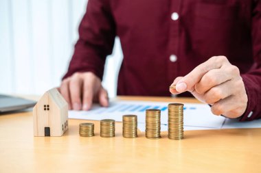 Asyalı işadamı madeni paralara zam yapmak için bozuk para tutuyor ev modeli ve evrak işlerinin mali tablosunu kontrol ederken emlak yatırımları ve ev kredisi için tasarruf hazırlığı yapıyor.