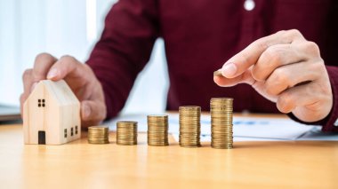 Asyalı işadamı madeni para tutarak ev modeline para yığıyor ve emlak yatırımı ve ev kredisi için tasarruf yaparken evrak işlerinin mali durumunu kontrol ediyor.