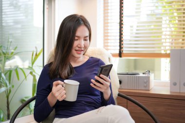 Genç Asyalı bir kadının arkadaşlarıyla sohbet etmek için akıllı telefon kullanması elinde kahve fincanı tutarken internetten alışveriş yapması ve oturma odasındaki rahat koltukta dinlenmesi.