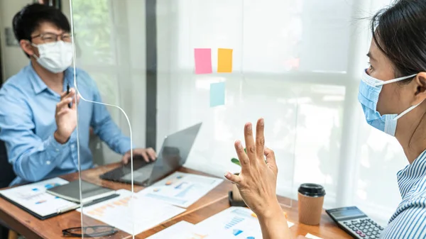 Close up of two business colleagues wearing face mask to brainstorming business project and giving high five to each other while working together with table shield partition to keep social distancing