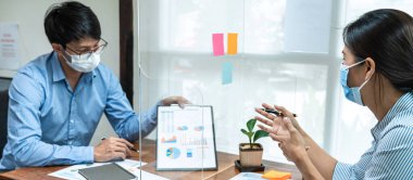 Meslektaşlarına finansal tabloyu gösteren Asyalı iş adamı ve yüz maskesi takıp sosyal mesafeyi korumak için masa kalkanı plastik bölme ile birlikte çalışırken yeni proje hakkında tartışan.