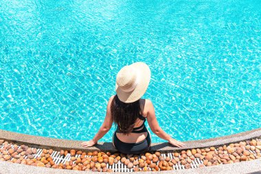 Asyalı bir yolcunun havuz kenarında oturup deniz manzarası seyrederken mayo ve güneş şapkası giymesinin keyfini çıkarması.