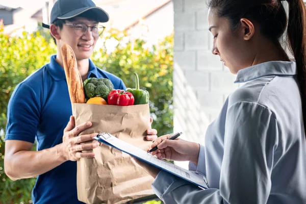 Asian woman customer order food delivery online and standing in front of the house to receive grocery of food, fruit, vegetable and sign on food delivery bill from courier