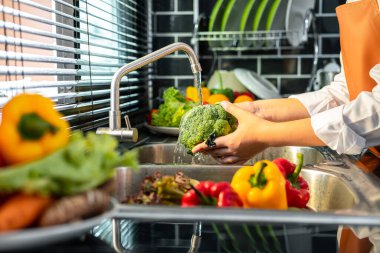 Asyalı ev hanımı önlük takıyor, brokoli ve sebzeleri yıkamak için lavaboda duruyor ve evde sağlıklı yemek pişirmek için lavaboya su koyuyor.